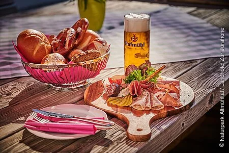 Ein gemütliche Brotzeit im Biergarten.
Hierzu wird ein Brotzeitbrettl mit einem Korb Brezen/Semmeln und einem Bier serviert.
