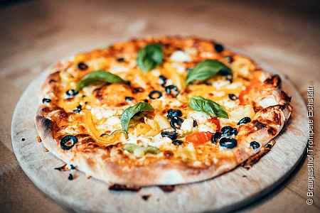 Eine Pizza mit fünf Basilikumblättern, Oliven und leckeren Käse liegt auf einem Holzbrett. Darunter sieht man das Holz eines Tisches.