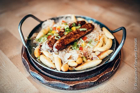 Zwei fränkische Bratwürste liegen wohl gebetet auf Schupfnudeln und Sauerkraut. Das Gericht ist in einem Eisenteller mit zwei Griffen angerichtet.