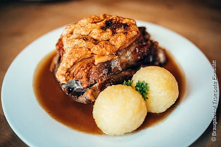 Auf einem weißen Teller sind zwei Kartoffelknödel mit Petersilie dazwischen angerichtet. Dahinter liegt das knusprige Schäuferl. Leckere Bratensoße zum Tunken füllt das weiße Teller aus.