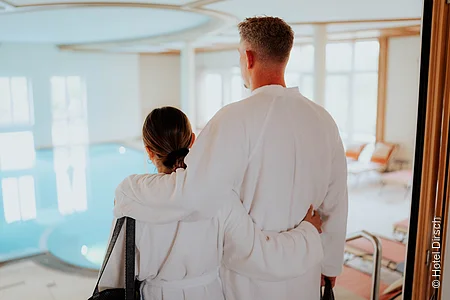 Ein Pärchen in weißen Bademäntel mit dem Rücken zu uns. Schauen auf den Indoorpool des Wellnessbereiches.