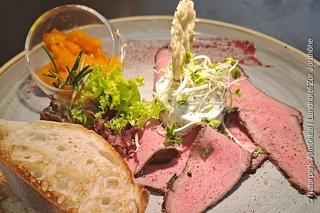 Teller mit Roastbeef, Salat, Brot, Frischkäse mit Sprossen und einem kleinen Glas mit orangener Sauce.